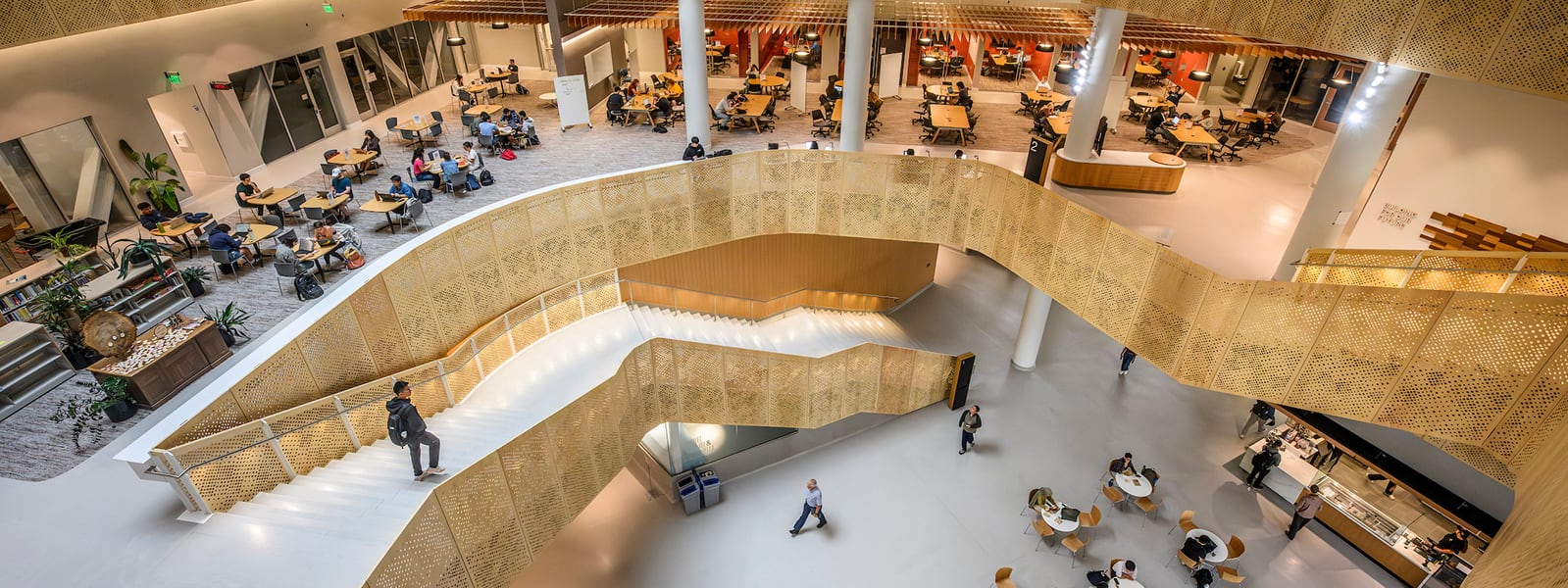 Interior view of Morgridge Hall -- a modern, multi-level building with curved golden staircase railings, open seating areas, and people walking and studying throughout the space.