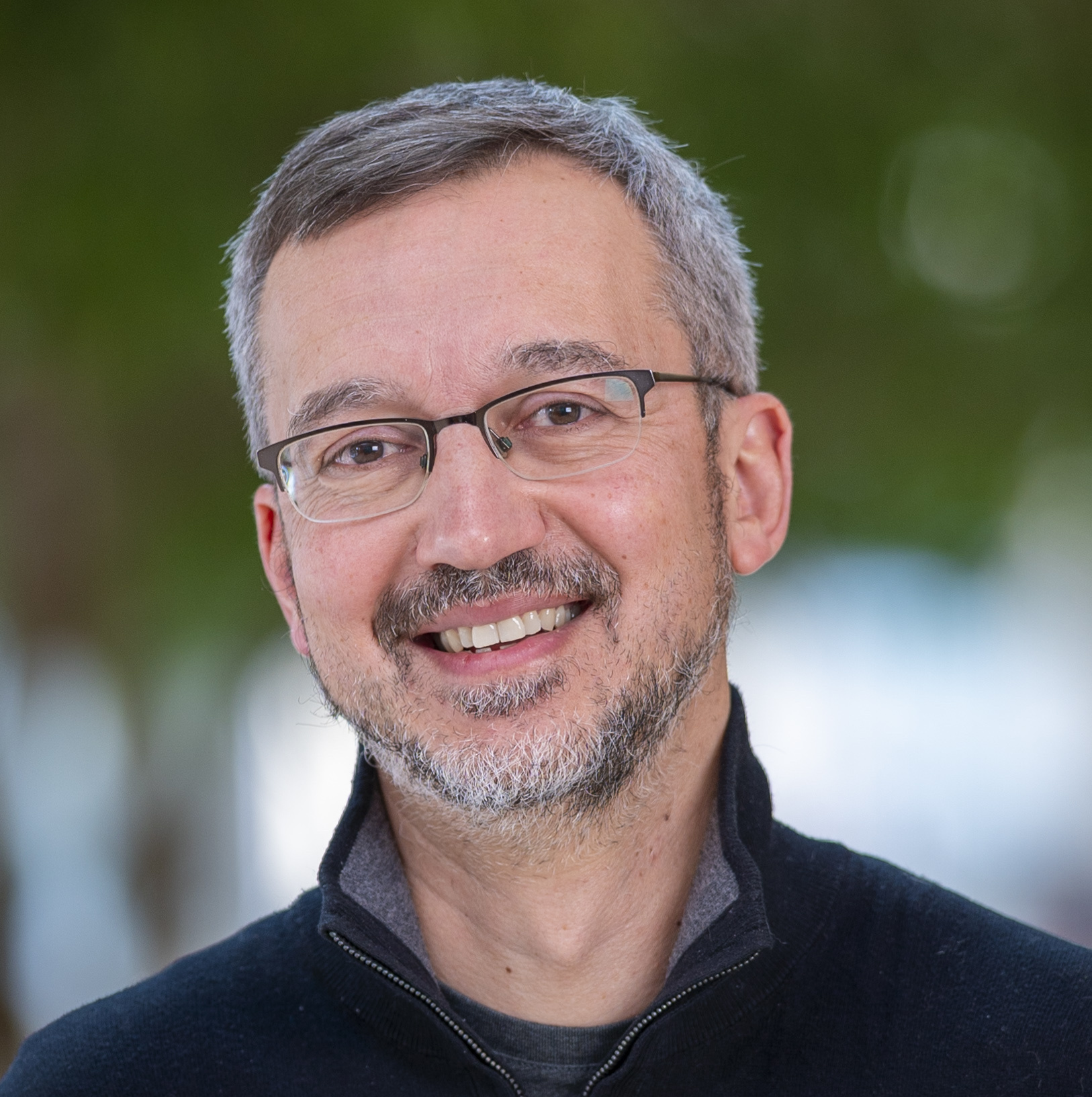 Remzi Arpaci-Dusseau wearing a black zip-up jacket with blurred green and blue background.