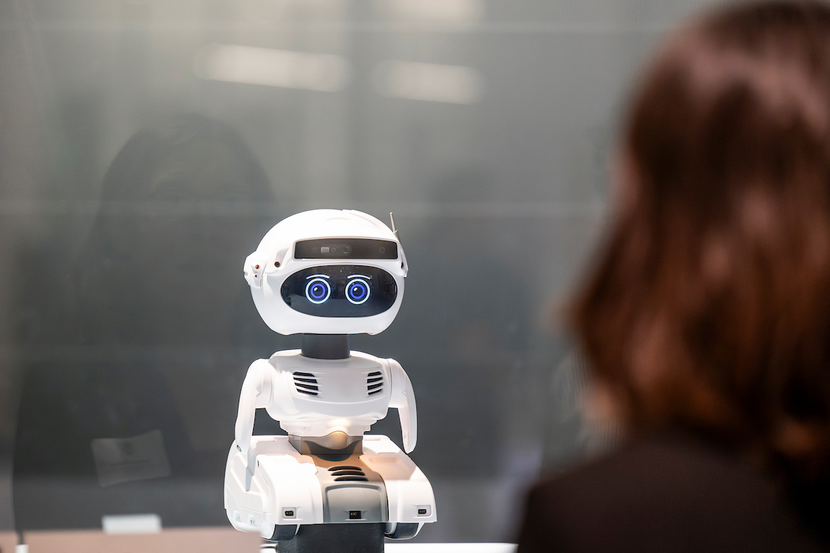Small white robot with glowing blue eyes facing a person across a glass barrier in a modern setting.