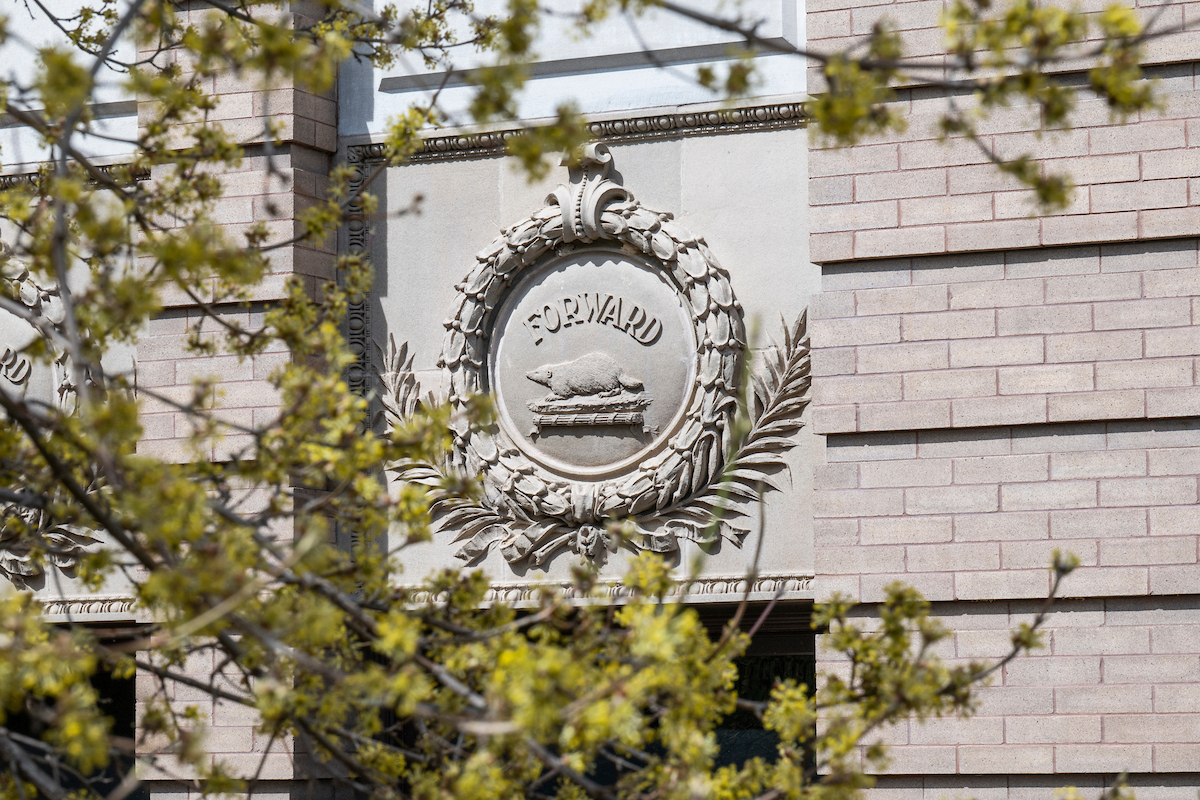 Stone emblem with the word "FORWARD" surrounded by a wreath on a building facade, partially obscured by tree branches.