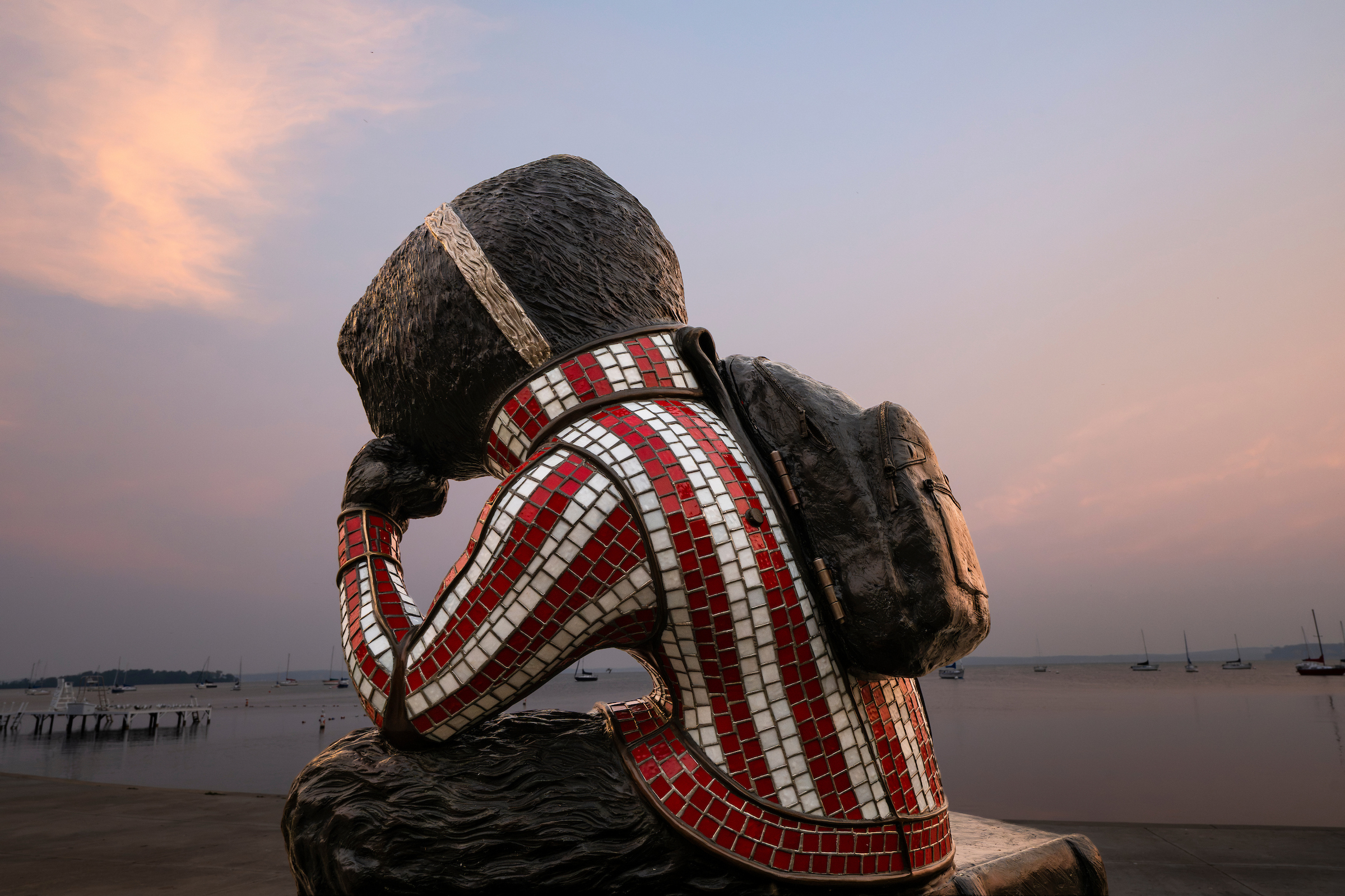 Sculpture of Bucky Badger wearing a red and white checkered shirt and backpack, sitting contemplatively by a calm waterfront at sunset.